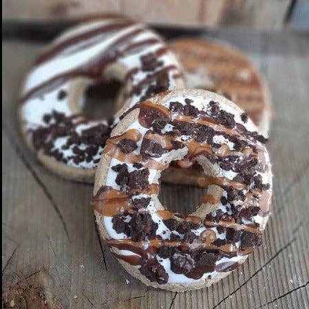 Three chocolate chip donuts with white icing, drizzles of chocolate and caramel sauce, and sprinkles, arranged on a rustic wooden surface.