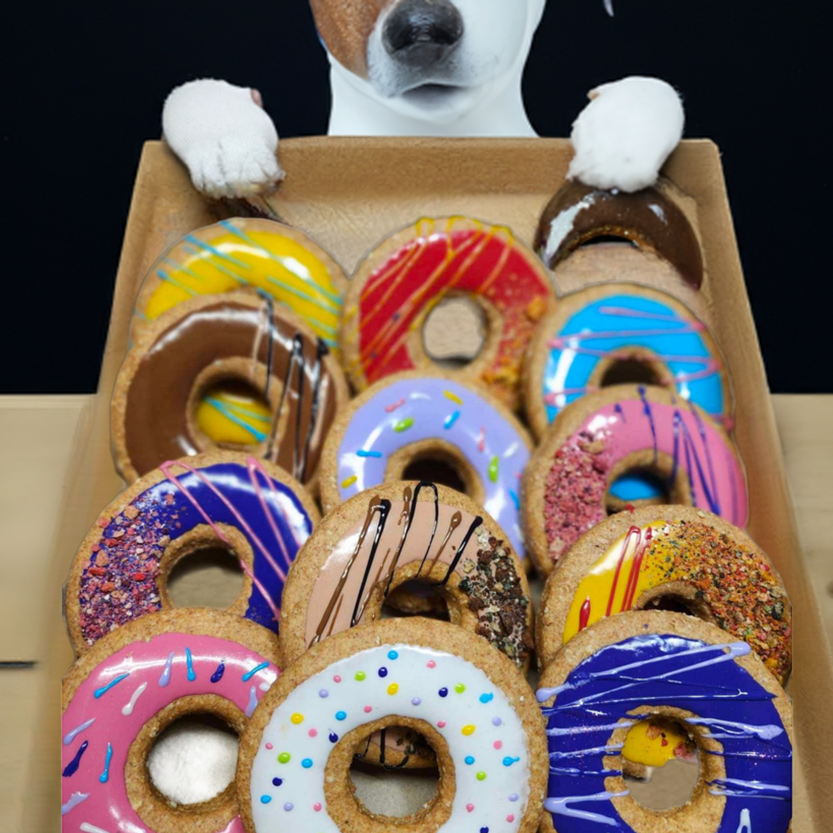 A cardboard tray filled with assorted colorful donuts, with a dog's paws resting on the tray and its head partially visible above it against a dark background.