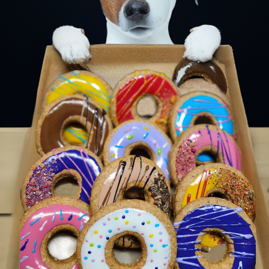 A cardboard tray filled with assorted colorful donuts, with a dog's paws resting on the tray and its head partially visible above it against a dark background.