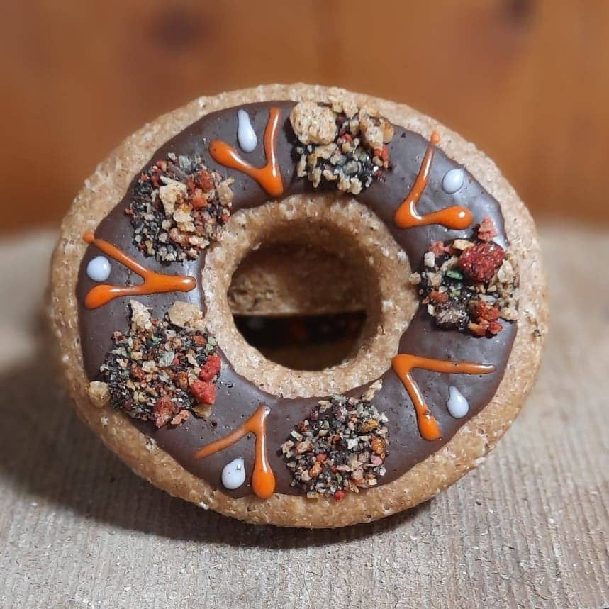 A novelty donut-shaped object with dark brown chocolate frosting, orange drizzle, and crumbled cookie toppings, photographed close-up on a textured beige surface with a blurred wooden background.