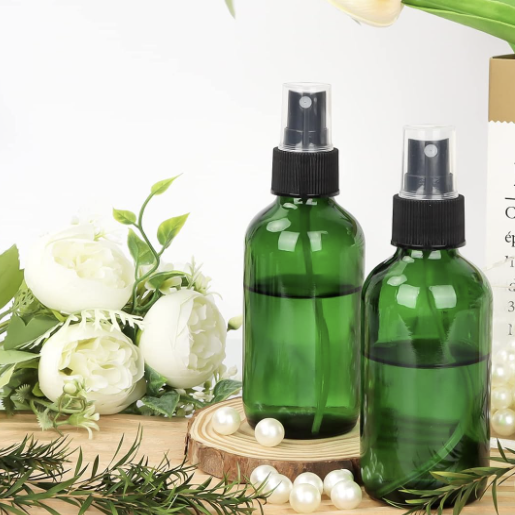 Two green glass spray bottles on a wooden surface with white flowers and greenery in the background.